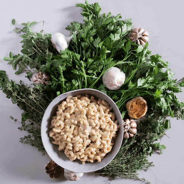 Bowl of vegan mac and cheese on top of a bed of herbs with bulbs of roasted garlic.